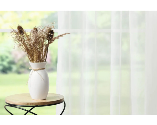 Fleurs séchées décoratives dans un vase blanc sur une petite table devant une fenêtre avec un rideau.
