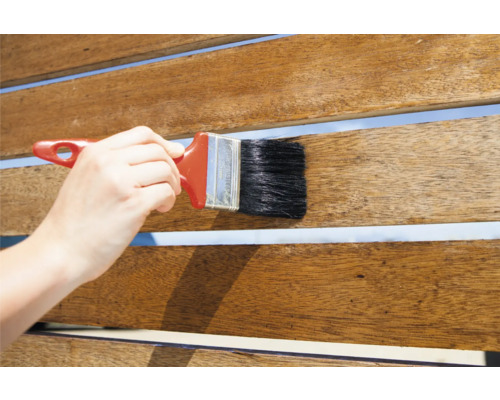 Eine Person trägt Holzlasur mit einem Pinsel auf ein Holzdeck auf.