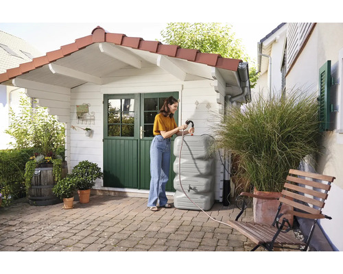 Une femme remplit un réservoir d''eau de pluie gris sur une terrasse devant un abri de jardin.