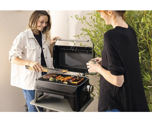 Deux femmes grillent des légumes et de la viande sur un barbecue portable à l''extérieur.