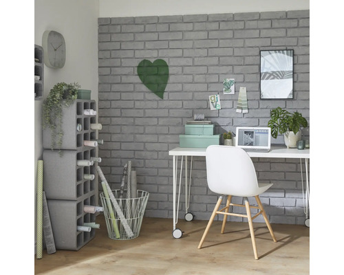 Arbeitszimmer mit Schreibtisch, Stuhl, Regalen und Dekorationsartikeln vor einer grauen Backsteinwand