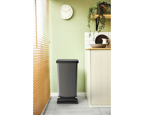 Poubelle à pédale grise dans la cuisine devant un placard de cuisine et un store vénitien