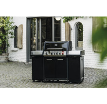 Schwarzer Gasgrill mit Unterschrank und Rädern im Außenbereich vor einem weißen Backsteingebäude.
