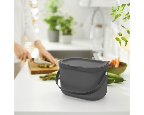 Poubelle avec couvercle sur un plan de travail de cuisine à côté de nourriture et d''une personne coupant des légumes.