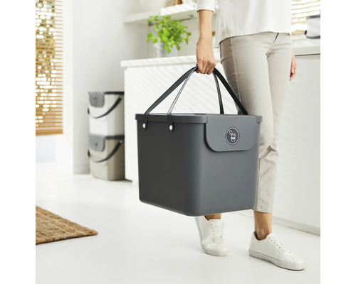 Une personne porte une poubelle grise avec des poignées et un symbole de recyclage du métal.