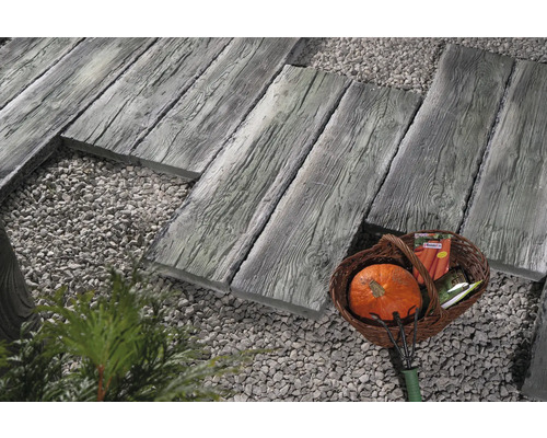 Gartenszene mit Gehwegplatten in Holzoptik, Zierkies und einem Korb mit Gartenutensilien.