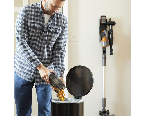 Un homme vide le réservoir à poussière d''un aspirateur sans fil dans une poubelle. Un chargeur mural avec un aspirateur se trouve sur le mur.