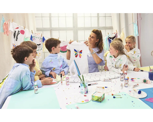 Gruppe von Kindern und eine Frau verzieren T-Shirts mit Textilfarbe an einem Tisch.