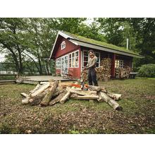 Mann mit Kettensäge beim Holzspalten vor einem Holzhaus mit Gründach.