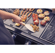 Préparation de nourriture sur un gril avec une spatule, des saucisses, des légumes, des tomates et des champignons farcis.