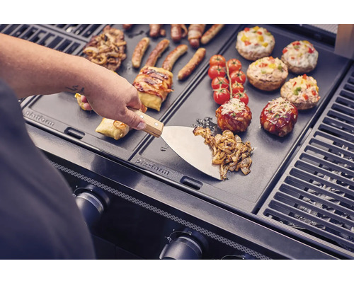 Préparation de nourriture sur un gril avec une spatule, des saucisses, des légumes, des tomates et des champignons farcis.