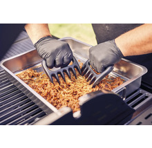 Zwei Hände mit Grillhandschuhen zerkleinern Fleisch mit Fleischkrallen in einer Metallschale auf einem Grill.
