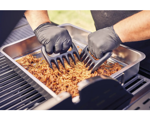 Zwei Hände mit Grillhandschuhen zerkleinern Fleisch mit Fleischkrallen in einer Metallschale auf einem Grill.
