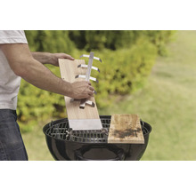 Szene mit Mann, der ein Grillbrett mit Halter über einem Holzkohlegrill hält, im Freien.