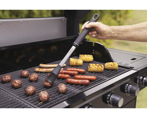 Scène de grillades avec brosse à gril, saucisses, épis de maïs et boulettes de viande sur un gril à gaz.