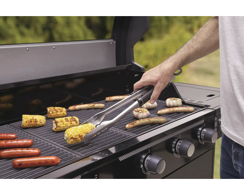 Grill mit Maiskolben, Würstchen und Knoblauch auf dem Grillrost und einer Person mit Grillzange.