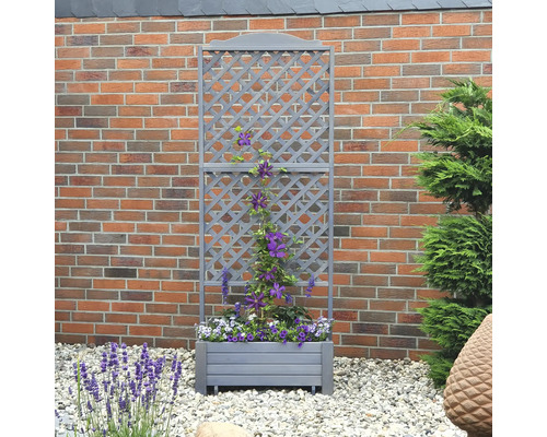 Jardinière grise avec treillis devant un mur de briques, plantée de fleurs violettes et blanches.