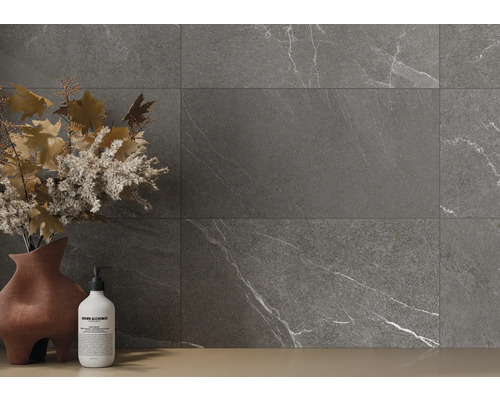 Graue Fliesen mit Marmoroptik an einer Wand, ergänzt durch eine Vase mit Blumenschmuck und eine Seifenflasche