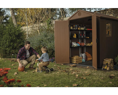 Scène montrant un abri de jardin en plastique avec des étagères, rempli de divers objets, tandis qu''un homme et un enfant jouent sur la pelouse.