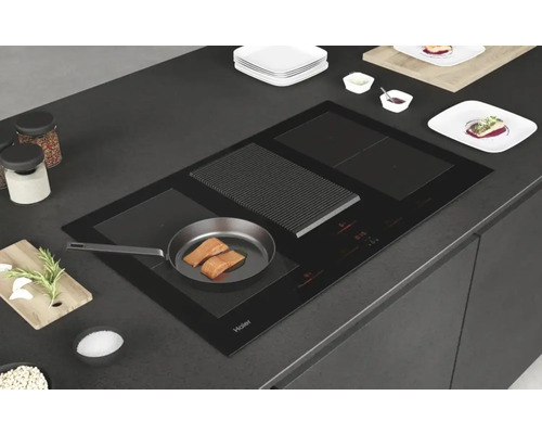 Plaque à induction noire Haier avec hotte intégrée, poêle avec poisson et ustensiles de cuisine sur un plan de travail