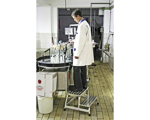 Mann in weißem Kittel steht auf einer Trittleiter und bedient eine Abfüllanlage für Flaschen