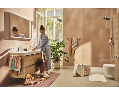 Helles Badezimmer mit Waschbecken, Spiegel, Dusche und Toilette. Eine Frau wäscht sich die Hände, während ein Kind auf dem Teppich spielt.