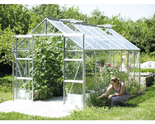 Gewächshaus mit geöffneten Fenstern und Pflanzen sowie einer Frau, die im Garten arbeitet.