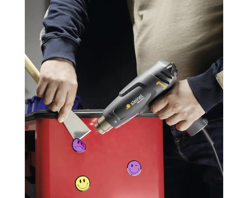 Une personne retire des autocollants d''une surface à l''aide d''un pistolet thermique et d''une spatule.