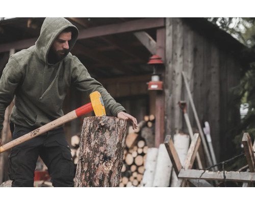 Un homme fend du bois avec un merlin devant une cabane en bois.