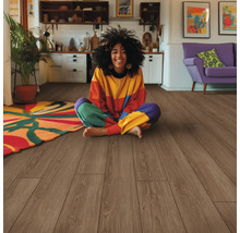 Une femme est assise sur un plancher en bois dans un salon lumineux avec une cuisine et un canapé à l''arrière-plan.