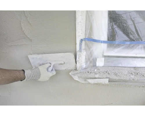 Handwerker glättet Putz an einer Wand mit einer Glättkelle neben einem Fenster, das mit Folie abgedeckt ist