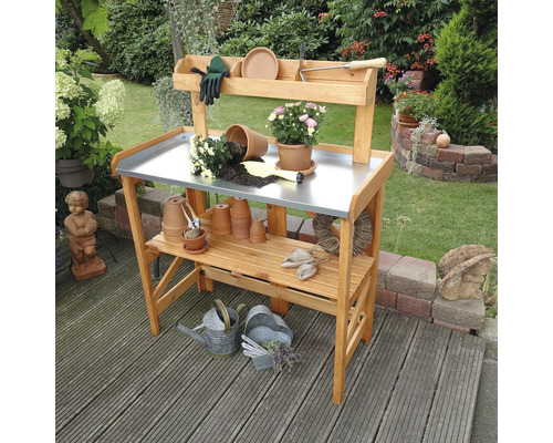 Table à rempoter rabattable avec tablette galvanisée 97 x 45 x 128 cm imprégnée Scène de jardin avec une table de rempotage en bois avec un plan de travail en métal et des étagères de rangement, entourée d''outils de jardin et de plantes.