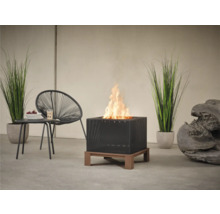 Quadratische Feuerstelle mit Flammen, Rattan-Design und Holzfuß auf einer Terrasse, ergänzt durch einen Stuhl und einen Tisch.