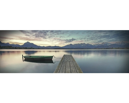 Ein ruhiger See mit einem Holzsteg und einem kleinen Boot vor einer Bergkulisse.