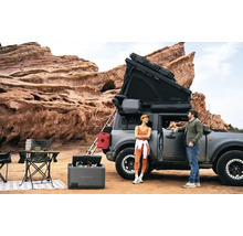 Szene mit einem Auto mit Dachzelt, einem tragbaren Kühlschrank, Campingmöbeln und zwei Personen in einer Outdoor-Umgebung.