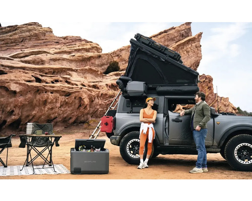 Szene mit einem Auto mit Dachzelt, einem tragbaren Kühlschrank, Campingmöbeln und zwei Personen in einer Outdoor-Umgebung.