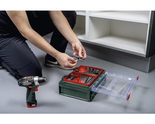 Eine Person hockt vor einem Regal und sortiert Bits aus einem Sortimentskasten, ein Akkuschrauber liegt bereit.