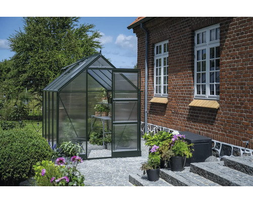 Gewächshaus mit geöffneter Tür in einem Garten neben einem Backsteingebäude.