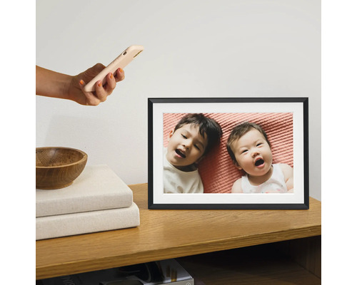 Digitaler Bilderrahmen auf einem Holzschrank mit einem Foto von zwei Kindern, die auf einem rosa Laken liegen. Eine Hand hält ein Smartphone darüber.