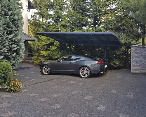 Ein graues Auto steht unter einem Carport auf einer gepflasterten Einfahrt.
