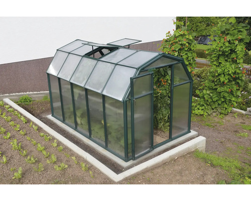 Grünes Gewächshaus in Scheunenform mit Dachfenstern und offener Tür in einem Garten auf Betonfundament.