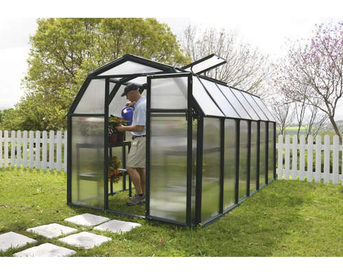 Gewächshaus mit schwarzem Rahmen und lichtdurchlässigen Paneelen im Garten. Ein Mann gießt Pflanzen in der hellen Struktur.