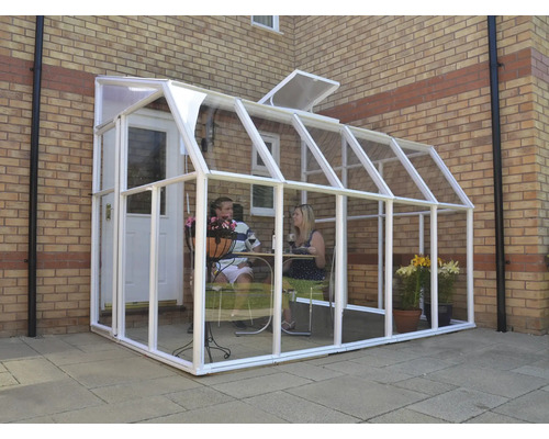 An eine Backsteinwand angelehntes Gewächshaus aus Glas mit weißem Rahmen, in dem zwei Personen an einem Tisch sitzen.