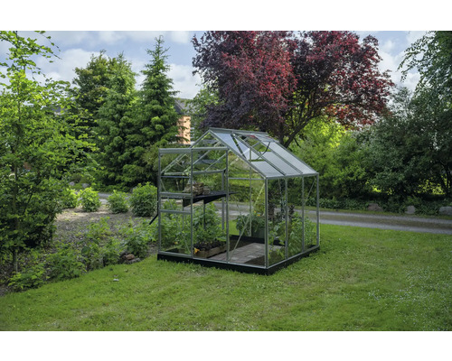 Serre transparente avec cadre en aluminium, fondation noire et lucarne sur une pelouse dans un jardin verdoyant.