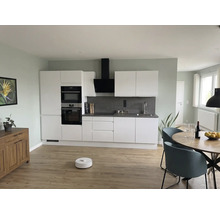 Cuisine moderne avec meubles blancs, crédence grise, table ronde en bois, chaises bleues et aspirateur robot sur un sol en bois clair.
