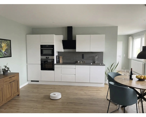Cuisine moderne avec meubles blancs, crédence grise, table ronde en bois, chaises bleues et aspirateur robot sur un sol en bois clair.