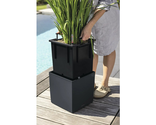 Personne soulevant un bac d insertion noir avec poignées d un pot de fleurs carré sur une terrasse en bois.