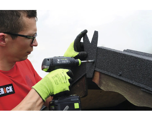Un homme utilise une perceuse visseuse sans fil pour fixer un élément de toiture en bitume sur une poutre en bois.
