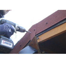 Une personne fixe des bardeaux de toiture en bitume avec une perceuse-visseuse à batterie sur une charpente en bois.
