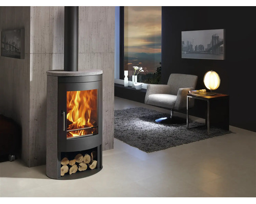 Moderner Kaminofen aus Stahl und Stein mit Feuer und Holzfach in hellem Wohnzimmer mit Sessel, Teppich und Betonoptik-Wand.